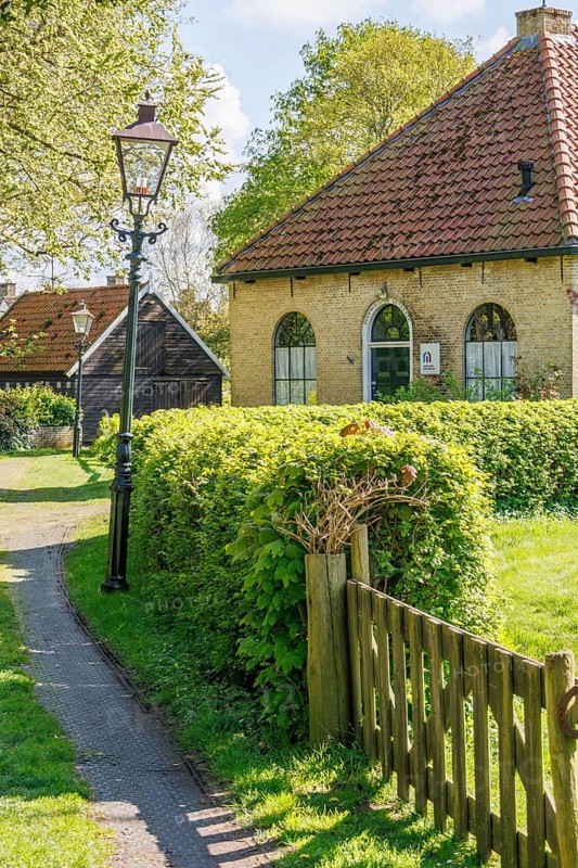 Traditional house in the village with surrounding meadow and hedges, illuminated by a lantern, trees in the background, historic houses in a small village with small streets and trees in spring, Nes, Ameland, Netherlands