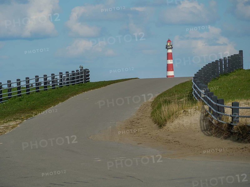 Road leads slightly uphill to a lighthouse, flanked by fences, with cloudy sky, red and white lighthouse by the sea, dunes, clouds and a cycle path, fence, nes, ameland, netherlands