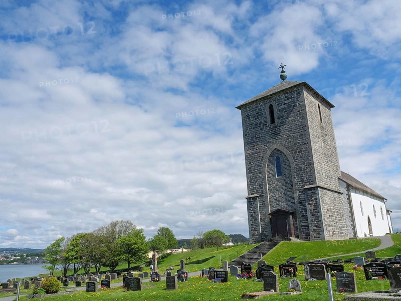 Church with cemetery under a cloudy sky on a green meadow, old stone church and many gravestones by the water with ships in the background, haugesund, norway
