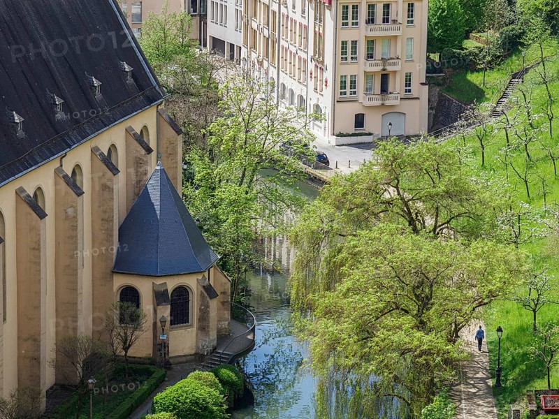 View of a church on the river bank, with neighbouring buildings and surrounded by lush greenery and trees, old church on a small river in the middle of a city against a cloudy sky, Luxembourg, Luxembourg, Europe