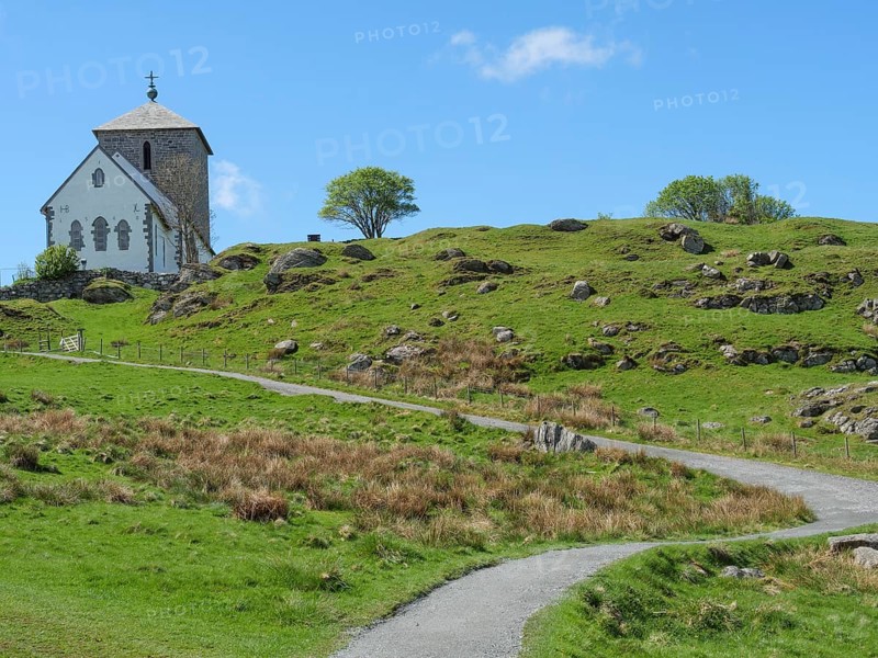 Church with hiking trail on hilly landscape, blue sky with clouds and trees, old stone church and many gravestones by the water with ships in the background, haugesund, norway