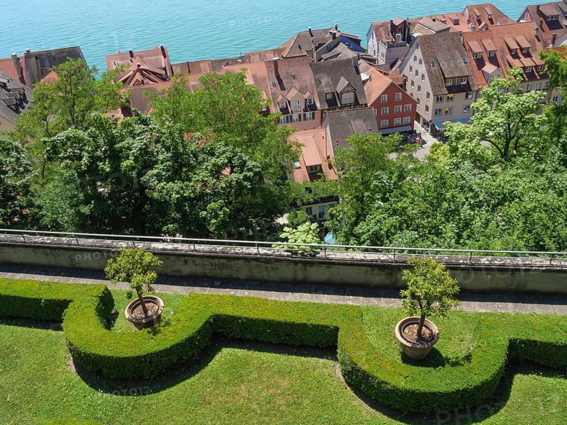 View of a picturesque garden with trees and trimmed hedges, in the background roofs and a lake, old buildings with many trees on a large lake, meersburg, germany