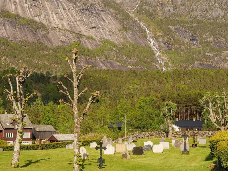 Cemetery with various graves and trees under a steep mountain landscape, gravestones with a small white church in front of a mountain panorama in spring, eidfjord, norway