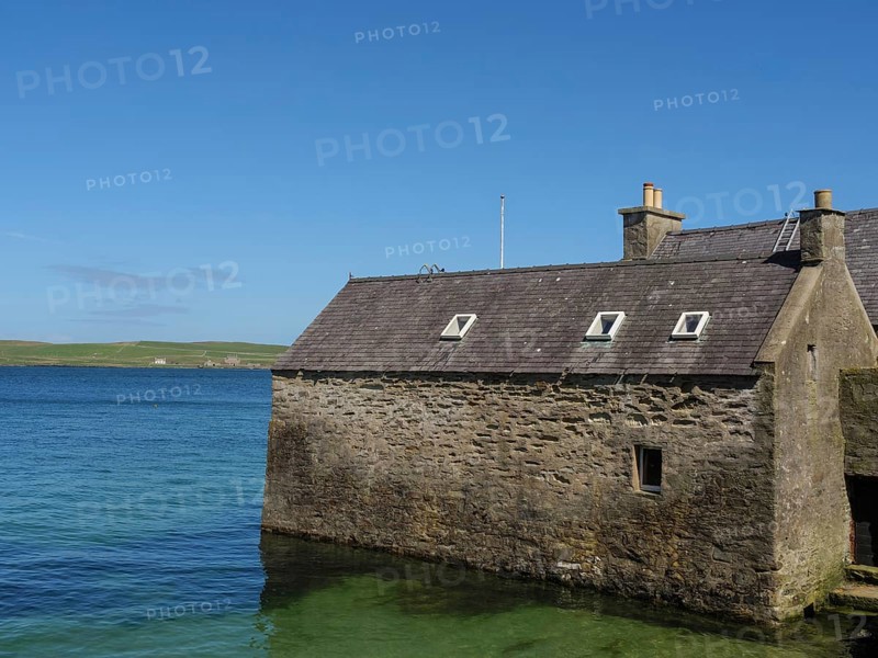 Small stone house on the shore of clear blue water with sweeping views of the coastline and blue sky, old stone house right on the blue sea in front of blue sky, lerwick, Scotland, United Kingdom, Europe