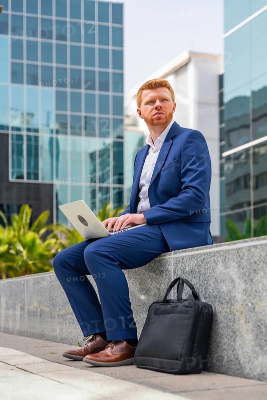 Vertical full length photo of a distracted businessman sitting outside a financial building using laptop