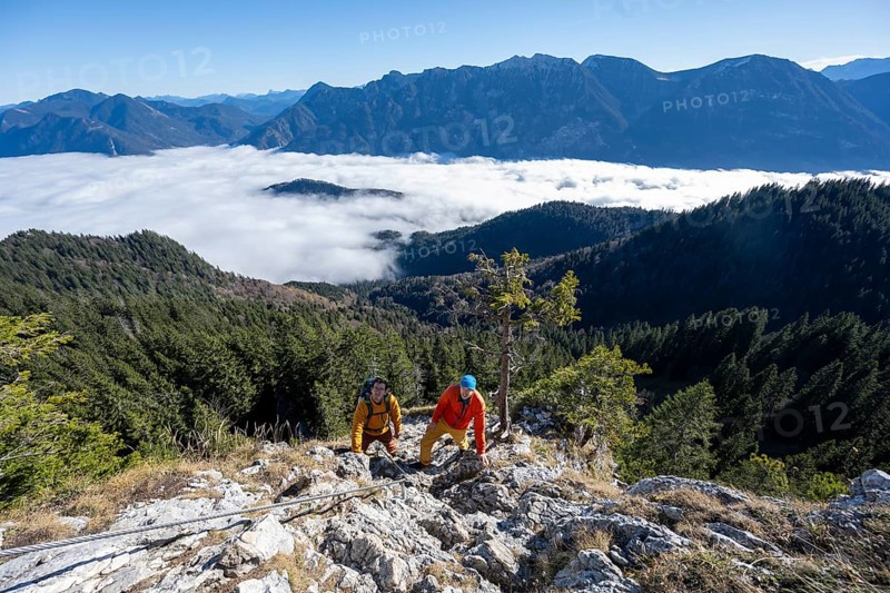 Two friends, mountaineers climbing the via ferrata to the rocky summit of the Ettaler Manndl, view over mountain landscape and sea of clouds, high fog in the valley, Ammergau Alps, Bavaria, Germany, Europe