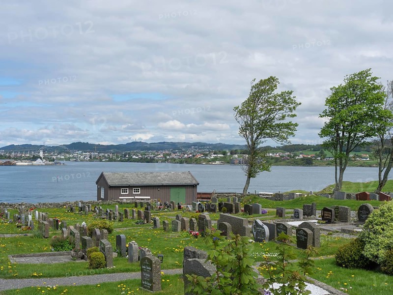 Cemetery with many gravestones, a small lake and a house under a partly cloudy sky, old stone church and many gravestones by the water with ships in the background, haugesund, norway