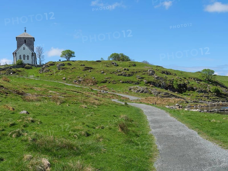 A small path leads to a church on a hill surrounded by green nature and a blue sky with few clouds, old stone church and many gravestones by the water with ships in the background, haugesund, norway