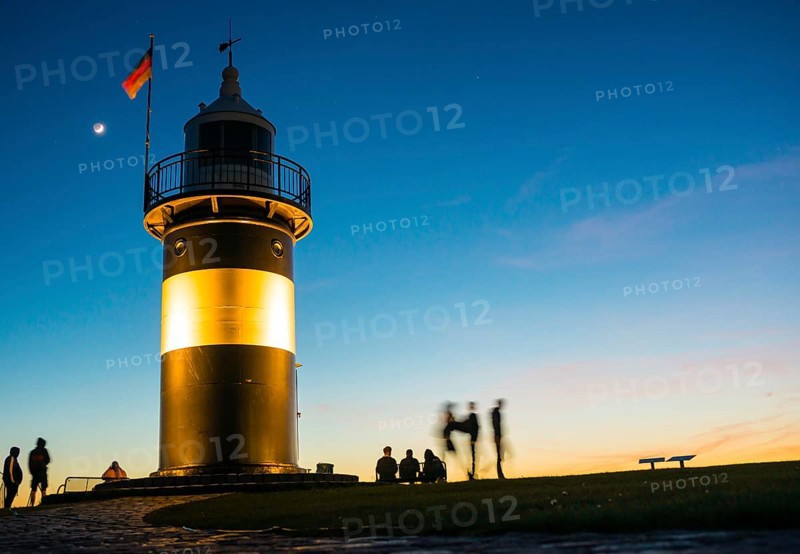 Black and white lighthouse, called 'Kleiner Preusse', stands illuminated at night shortly after sunset at the end of a paved, curved path, silhouettes of people next to it, wipe effect, path in the foreground, the waxing moon is in the blue sky, Crescent with visible dark moon disc, Germany flag, evening, close-up, North Sea resort of Wremen, Lower Saxony Wadden Sea National Park, World Heritage Site, Wurster North Sea coast, Land Wursten, Cuxhaven district, Lower Saxony, Germany, Europe