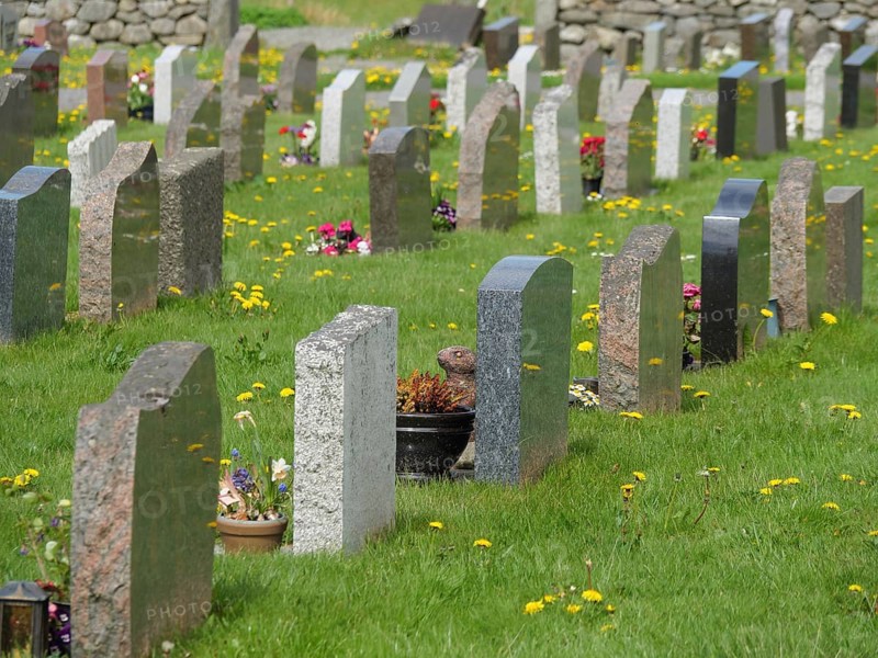 Row of gravestones on a green lawn with spring flowers in a quiet environment, old stone church and many gravestones by the water with ships in the background, haugesund, norway