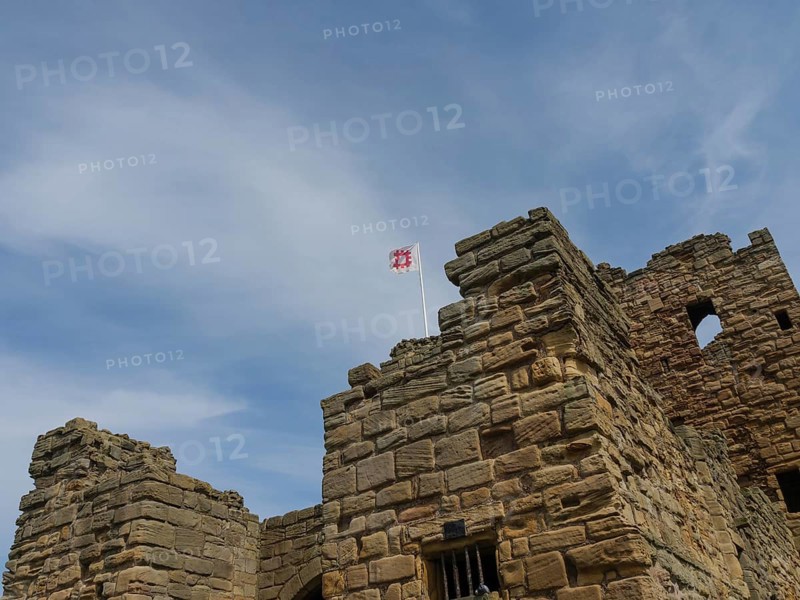 The top of a historic castle with stone towers and a waving flag under a cloudy sky, ruins and old stone walls by the sea, green fields with ruins, Newcastle, England, United Kingdom, Europe