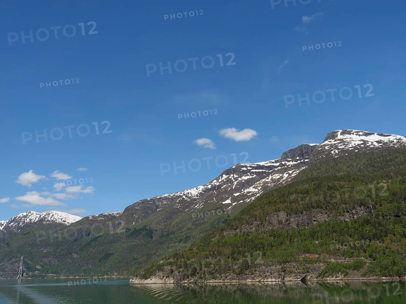 A clear day with snow-capped mountains under a blue sky with scattered clouds, bridge in a fjord with snow-capped mountains and green trees, greenish shimmering water, eidfjord, Norway, Europe