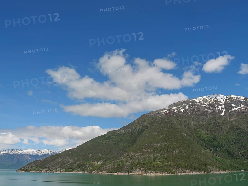 A mountain under a clear blue sky with some clouds, surrounded by green forests and a lake, greenish shimmering water in a fjord with snow-capped mountains and green trees, eidfjord, Norway, Europe