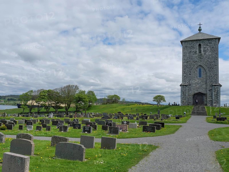 Stone church in a cemetery with numerous gravestones in a quiet, rural setting, old stone church and many gravestones by the water with ships in the background, haugesund, norway