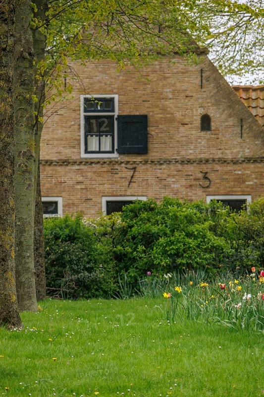 Brick house with black shutters and numbered facade, surrounded by a green meadow and flowers, old houses with green gardens in a small village, nes, ameland, the netherlands