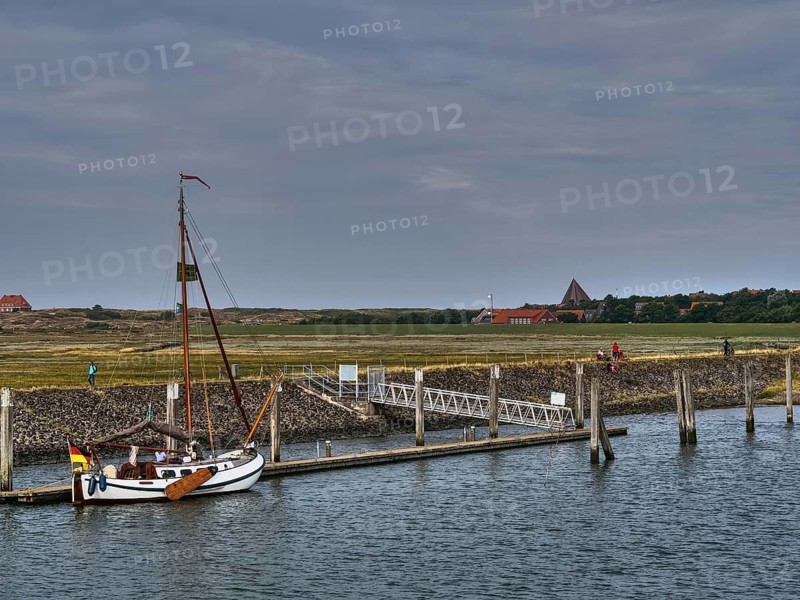 A sailing boat is moored at a pier, houses and people can be seen in the background, all under a cloudy sky, small jetty with a one-sea sailing boat in the north sea in front of a cloudy sky, spiekeroog, germany