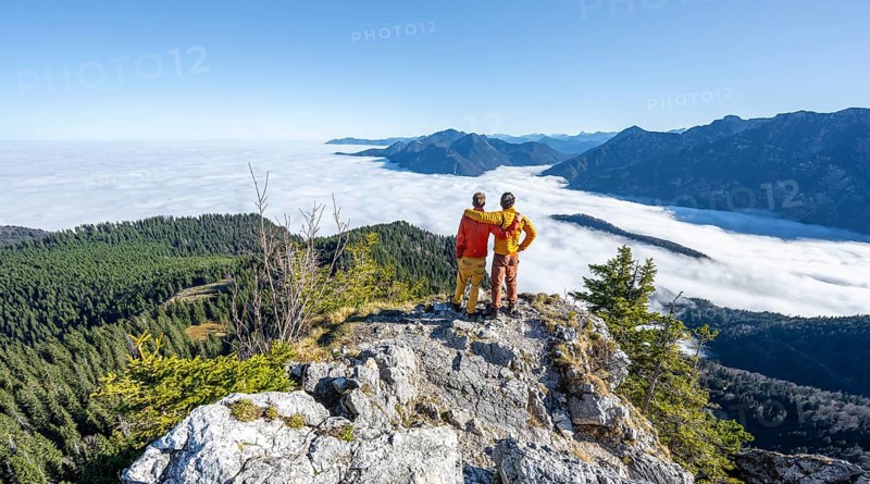 Two friends put their arms around each other's shoulders, mountaineers on the rocky summit of the Ettaler Manndl, view over mountain landscape and sea of clouds, high fog in the valley, Ammergau Alps, Bavaria, Germany, Europe