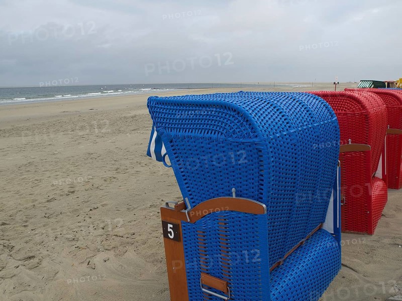 A blue beach chair stands on the beach with other red beach chairs in the background under a cloudy sky, beach chairs and beach tents by the sea under a cloudy sky, boardwalk in the sand, Borkum, Germany, Europe