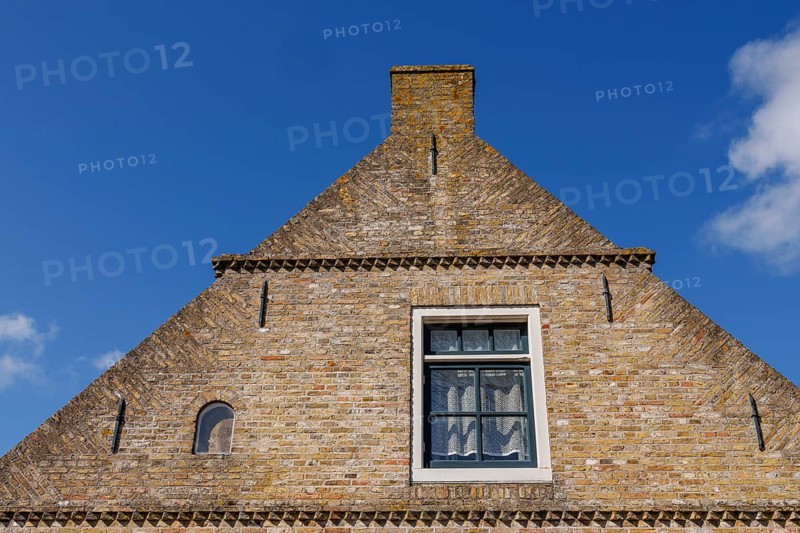 Close-up of a rustic brick house gable with a window under a bright blue sky, historic houses and a church with small streets and lanterns, Nes, Ameland, Netherlands