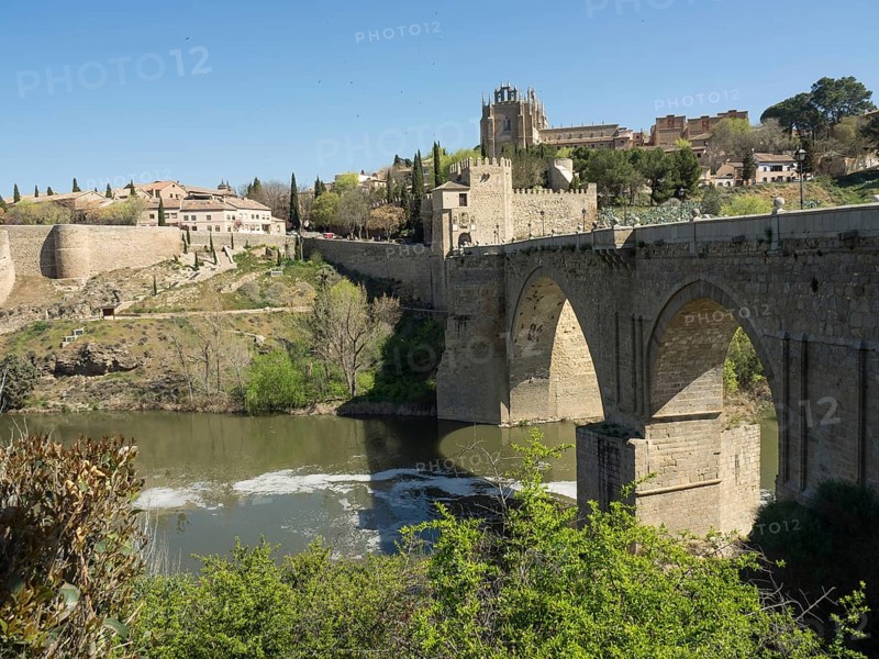 Stone bridge with arches over a river, surrounded by a cityscape and vegetation, sunny weather, historic stone bridge with an old tower and an old town in the background over a wild river, Toledo, Spain, Europe