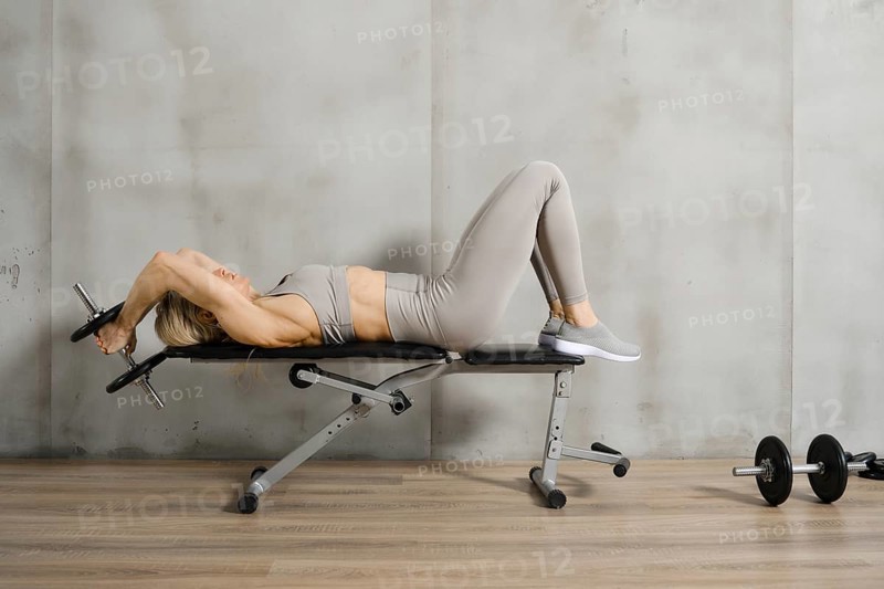 A muscular woman lies on a bench in the gym and pushes a dumbbell behind her head with both hands