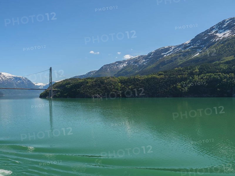 Turquoise lake surrounded by snow-capped mountains, a bridge spans in the background under a blue sky, bridge in a fjord with snow-capped mountains and green trees, greenish shimmering water, eidfjord, Norway, Europe
