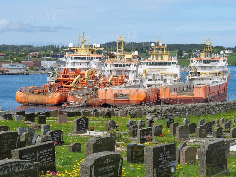 Cemetery with gravestones and several ships in the background under a slightly cloudy sky, old stone church and many gravestones by the water with ships in the background, haugesund, norway