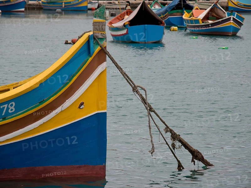 A yellow-blue boat is moored to the harbour with a rope, surrounded by other boats, many colourful fishing boats in a harbour in the Mediterranean, Marsaxlokk, malta