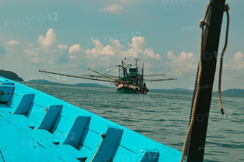 A fishing boat is seen on calm ocean waters under a cloudy sky from the bow of a blue boat, showing a candid moment of daily life and livelihood in the island of Koh Sdach in Cambodia