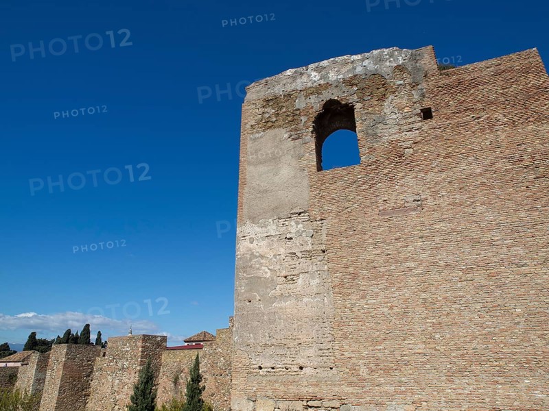 High stone tower with window opening surrounded by blue sky and sunlight, stone walls of an old Moorish style fortress with palm trees and blossom against blue sky, malaga, spain