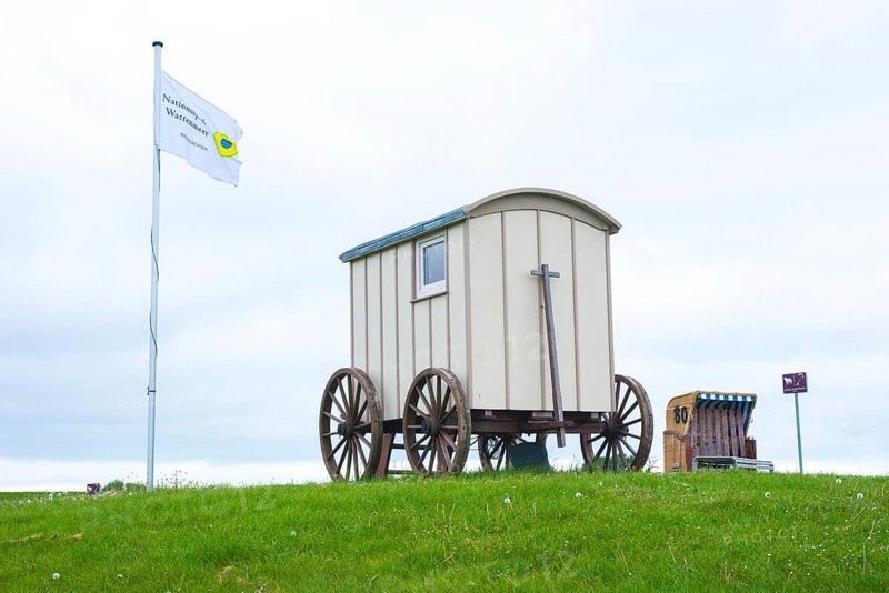 Old wooden cart, bathing cart with drawbar and large wooden wheels on a green meadow with a flag next to it, in the background a beach chair, cloudy day, deserted, sea, flag blowing in the wind, North Sea, North Sea resort Wremen, Lower Saxony Wadden Sea National Park, World Heritage Site, Wurster North Sea coast, Land Wursten, district of Cuxhaven, Lower Saxony, Germany, Europe