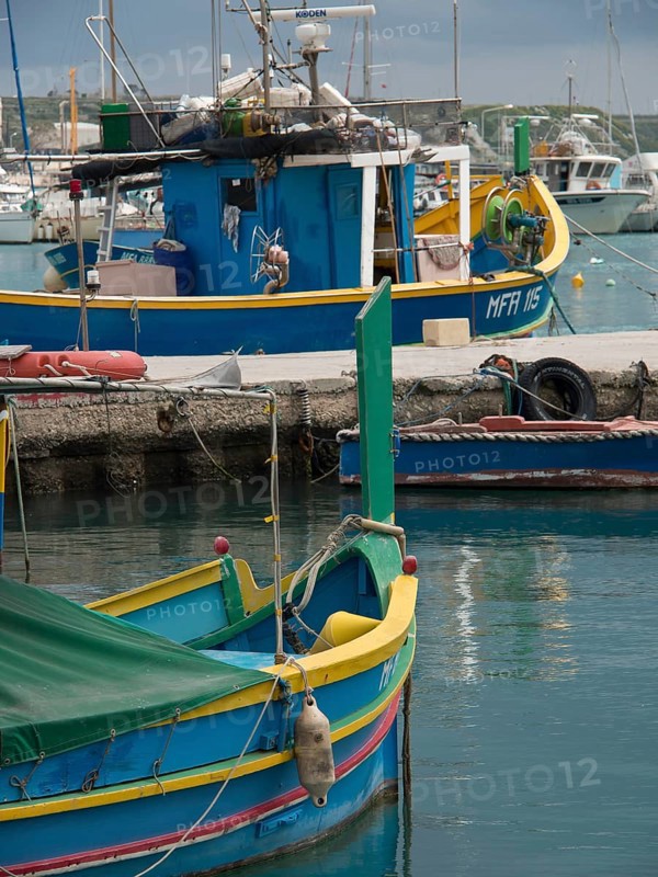 Small colourful boats moored in the water with other boats and huts in the background, many colourful fishing boats in a harbour in the Mediterranean, Marsaxlokk, malta