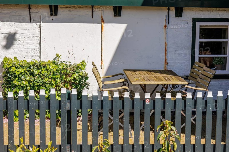 Garden furniture made of wood and metal behind a fence, with plants next to a sunlit wall, historic houses and a church with small streets and lanterns, Nes, Ameland, Netherlands