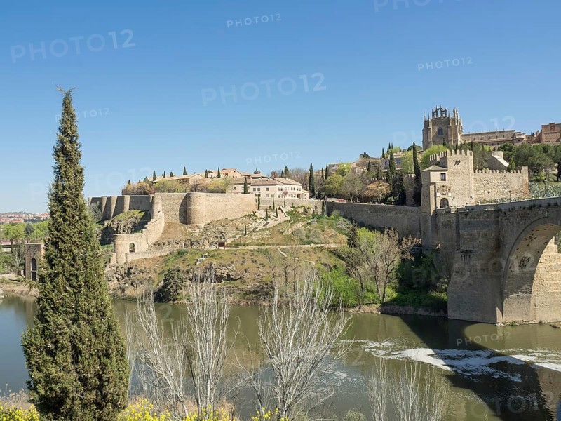 Historic fortress with walls and cypress trees on a hill with river in the foreground on a sunny day, historic stone bridge with an old tower and an old town in the background over a wild river, Toledo, Spain, Europe