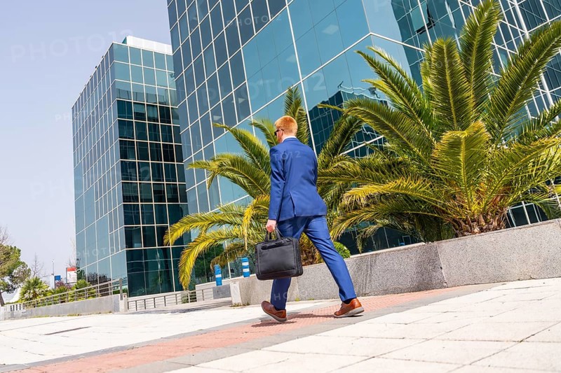 Horizontal full length photo of the rear view of a businessman walking holding a laptop bag along a financial district