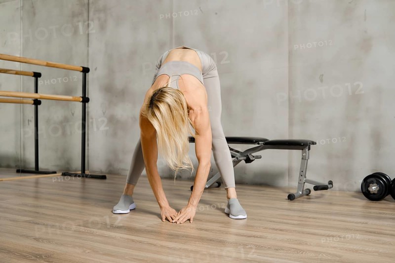 Middle-aged woman stretching back and arms in gym. Fitness athlete does bents and touching floor with her palms