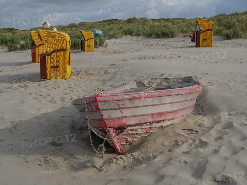 An abandoned boat half sunk in the sand, surrounded by beach chairs and dunes, colourful beach chairs on the beach and in the dunes under cloudy sky, red wooden boat and yellow beach chair, Juist, Germany, Europe