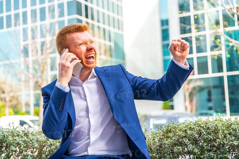 Businessman raising fist celebrating while talking to the mobile sitting in the city