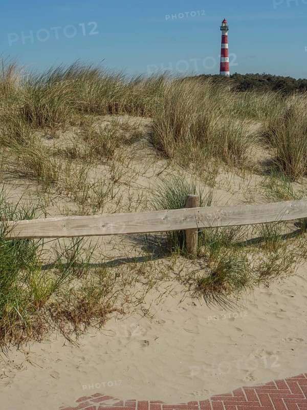 A lighthouse stands behind sandy dunes and grasses with a clear blue sky, red white lighthouse by the sea, dunes, clouds and a cycle path, fence, nes, ameland, netherlands