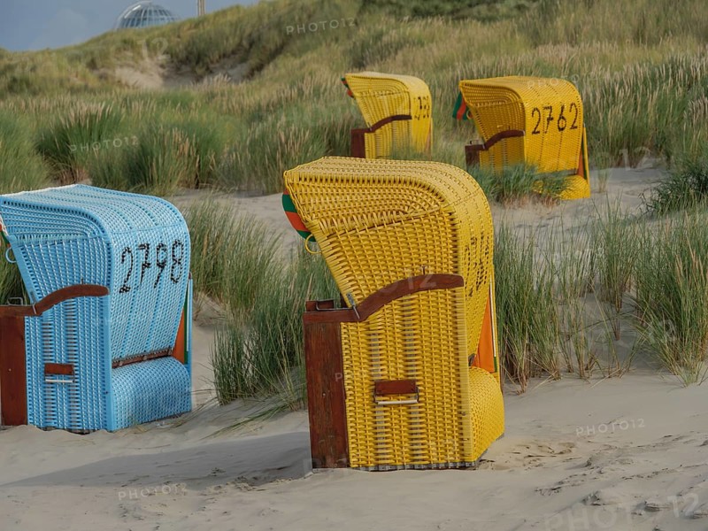 Colourful beach chairs standing on the sandy beach, surrounded by green dunes and a cloudy sky, colourful beach chairs on the beach and in the dunes under a cloudy sky, red wooden boat and yellow beach chair, Juist, Germany, Europe