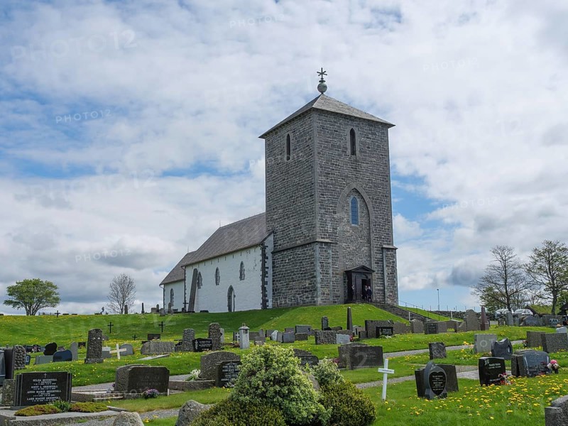 Stone church on a hill surrounded by a cemetery with numerous gravestones under a cloudy sky, old stone church and many gravestones by the water with ships in the background, haugesund, norway