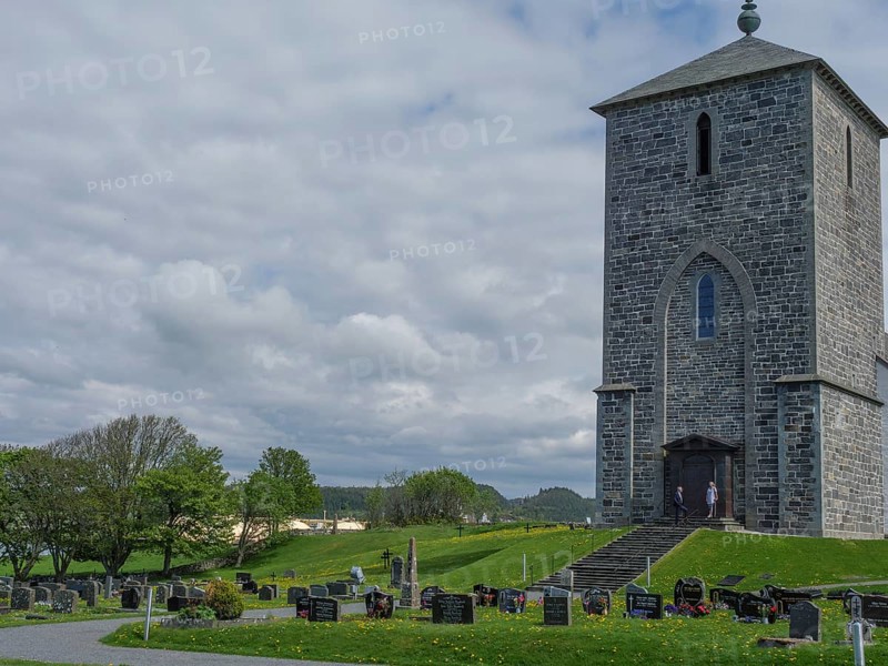 Stone church and cemetery with numerous gravestones under a cloudy sky in a rural setting, old stone church and many gravestones by the water with ships in the background, haugesund, norway