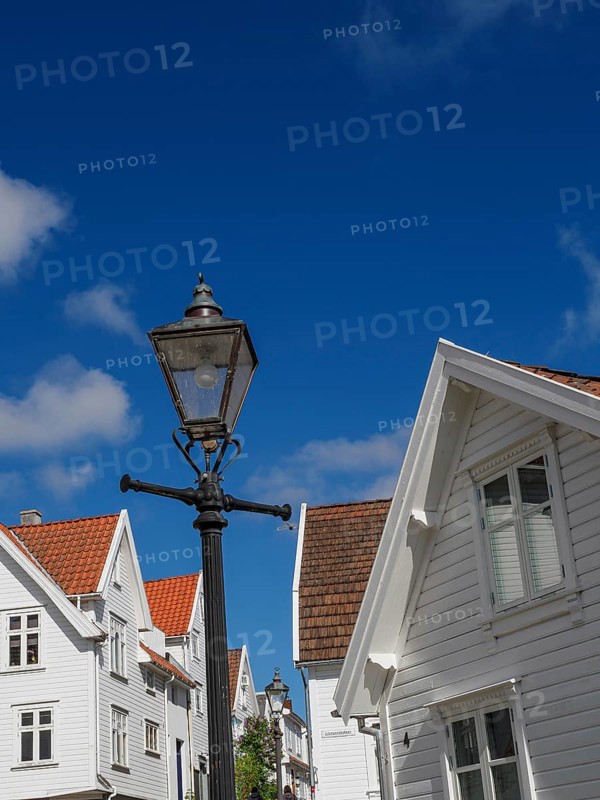 Street lamp in front of white houses with red roofs under blue sky with clouds, white wooden houses with red roofs in front of blue sky, trees and white fences on the street, stavanger, norway