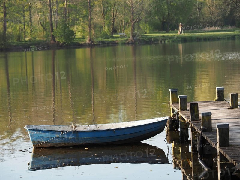 A boat is moored to a jetty and reflected in the calm waters of a lake, rowing boats and pedal boats on a small wooden jetty in the lake surrounded by trees, Borken Germany