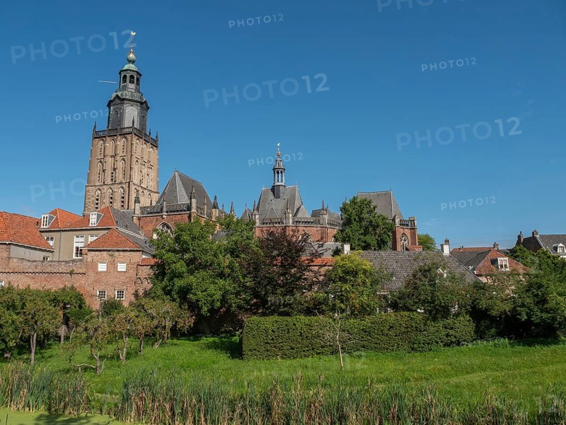 Historic church with a bell tower, surrounded by brick buildings and a green park under a clear midday sky, church tower and historic houses with trees, zutphen, Netherlands