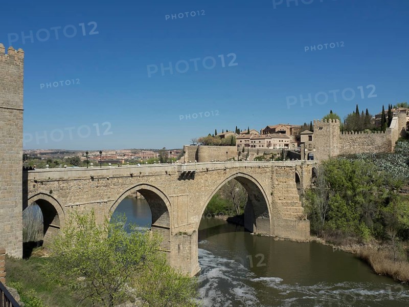 Historic stone bridge over a river surrounded by a city and nature, blue sky in the background, historic stone bridge with an old tower and an old city in the background over a wild river, Toledo, Spain, Europe
