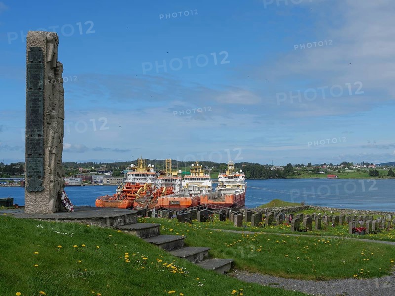 Stone memorial in a cemetery with a view of the harbour and several anchored ships, old stone church and many gravestones on the waterfront with ships in the background, haugesund, norway