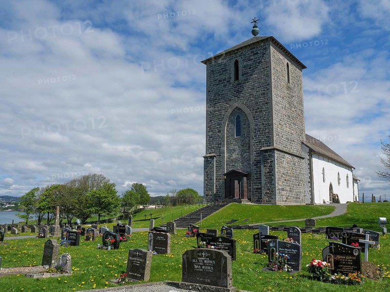 Old church on a cemetery under a blue cloudy sky, old stone church and many gravestones on the water with ships in the background, haugesund, norway