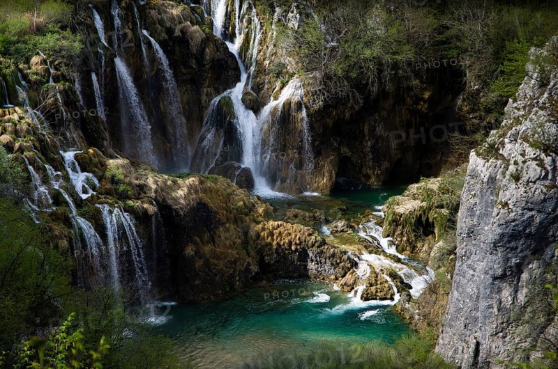 A picturesque waterfall that pours over rocks into crystal-clear water, surrounded by lush green vegetation and dense forests. Plitvice Lakes National Park, Croatia, Europe