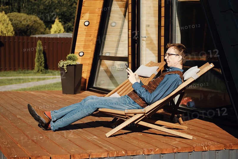 Middle aged man using smartphone while resting on terrace of wooden cabin in a sunny day